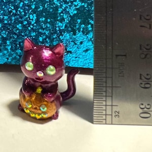 May include: A small, metallic magenta cat figurine holding an orange pumpkin with a carved face. The cat has iridescent green eyes and a rhinestone nose. A metal ruler is positioned next to the figurine, showing a height of approximately 1 inch.