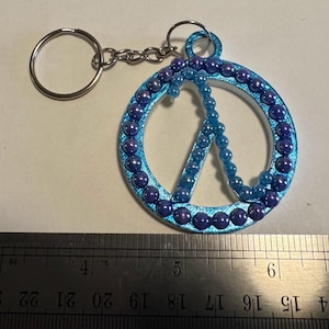 May include: A blue peace sign keychain with a silver key ring and chain. The peace sign is decorated with blue and purple beads around the edge and in the center. The keychain is on a white surface.