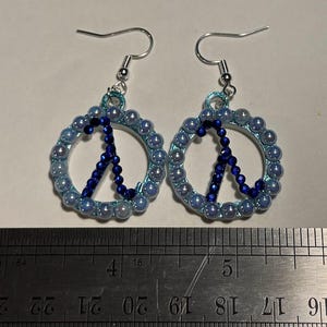 Op de afbeelding: Een paar oorbellen met een vredestekenontwerp. Elke oorbel heeft een lichtblauwe kralencirkel met een donkerblauw kralen vredessymbool in het midden. De oorbellen hebben zilverkleurige oorhaken.