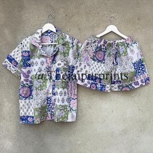 Peut inclure: Un ensemble de pyjama deux pièces avec un motif patchwork floral. L'ensemble comprend une chemise boutonnée à manches courtes et un short assorti. La palette de couleurs présente des teintes blanches, bleues, vertes et roses. L'ensemble est suspendu à des cintres blancs.