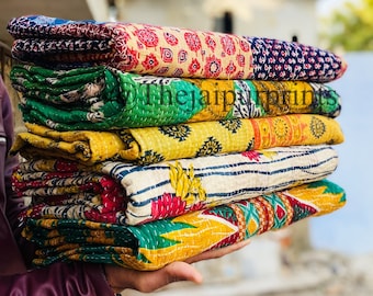 May include: A stack of six colorful kantha quilts, folded and arranged in a person's hand. The quilts are made of patchwork fabric with various patterns and colors, including red, yellow, green, blue, and white.
