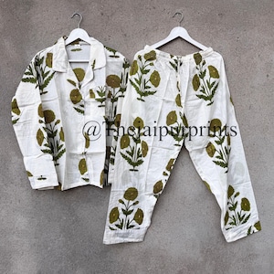 May include: White pajama set with a green floral print. The set includes a button-down shirt and matching pants, both hanging on a white hanger. The floral design is repeated throughout the shirt and pants.