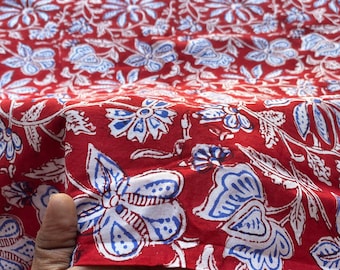 May include: Red cotton fabric with a white and blue floral print. The fabric is folded in half and a hand is holding the fabric.