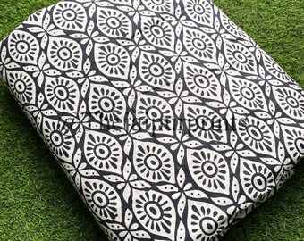May include: Folded fabric with a repeating black and white floral pattern. The design features stylized flowers and geometric shapes. The fabric is resting on a green surface, suggesting a textile or craft item.