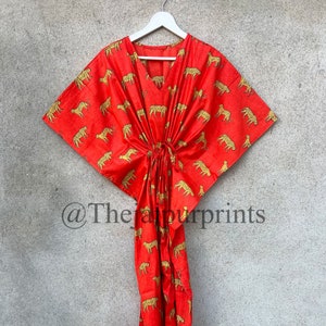 May include: A red kaftan with a leopard print design. The kaftan has a V-neck and is tied at the waist. The kaftan is hanging on a white hanger against a gray wall. The text "@TheJaipurprints" is visible in the bottom left corner of the image.