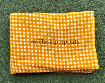 May include: Folded fabric with a yellow and white checkered pattern. The fabric is a warm yellow color with a grid of white squares. The text "@thejaipurprints" is visible on the fabric. The fabric is lying on a green surface.