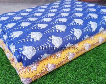 May include: Three pieces of fabric with floral patterns. The top fabric is blue with white flowers, the middle fabric is yellow with white flowers, and the bottom fabric is yellow with red flowers. The fabrics are folded and stacked on top of each other. The fabrics are on a green grassy surface.