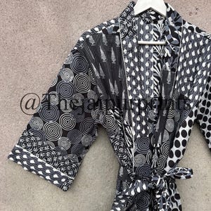 May include: A black and white patterned robe with a tie belt, hanging on a white hanger. The robe features various geometric and floral designs in shades of gray and white. The robe's edges are trimmed with a white border.
