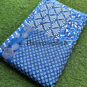 以下が含まれることがあります： 青と白の綿生地でできたパッチワークのデザイン。生地には、円、渦巻き、葉など、さまざまなパターンが施されています。生地は半分に折り畳まれ、緑色の表面の上に置かれています。