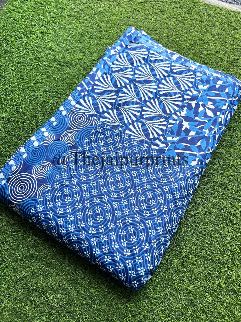 以下が含まれることがあります： 青と白の綿生地でできたパッチワークのデザイン。生地には、円、渦巻き、葉など、さまざまなパターンが施されています。生地は半分に折り畳まれ、緑色の表面の上に置かれています。