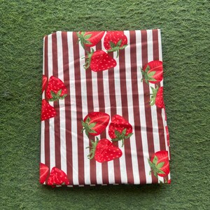 May include: Folded fabric with a pattern of red strawberries and green leaves on a white and brown striped background. The fabric is rectangular and appears to be folded neatly. The strawberries are evenly spaced across the fabric.