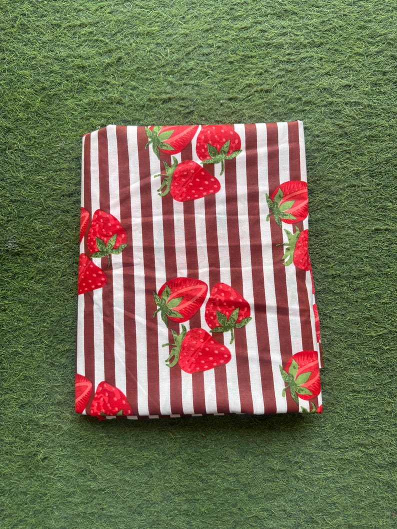 May include: Folded fabric with a pattern of red strawberries and green leaves on a white and brown striped background. The fabric is rectangular and appears to be folded neatly. The strawberries are evenly spaced across the fabric.