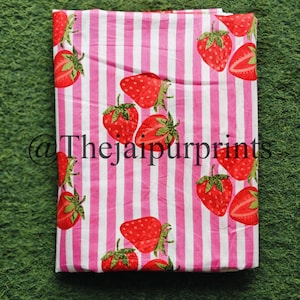 May include: A folded fabric featuring a repeating pattern of red strawberries with green leaves on a pink and white striped background. The text "@thejaipurprints" is visible in the lower left corner. The fabric is likely cotton or linen.