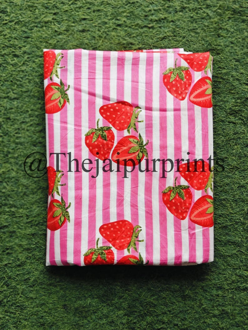 May include: A folded fabric featuring a repeating pattern of red strawberries with green leaves on a pink and white striped background. The text "@thejaipurprints" is visible in the lower left corner. The fabric is likely cotton or linen.