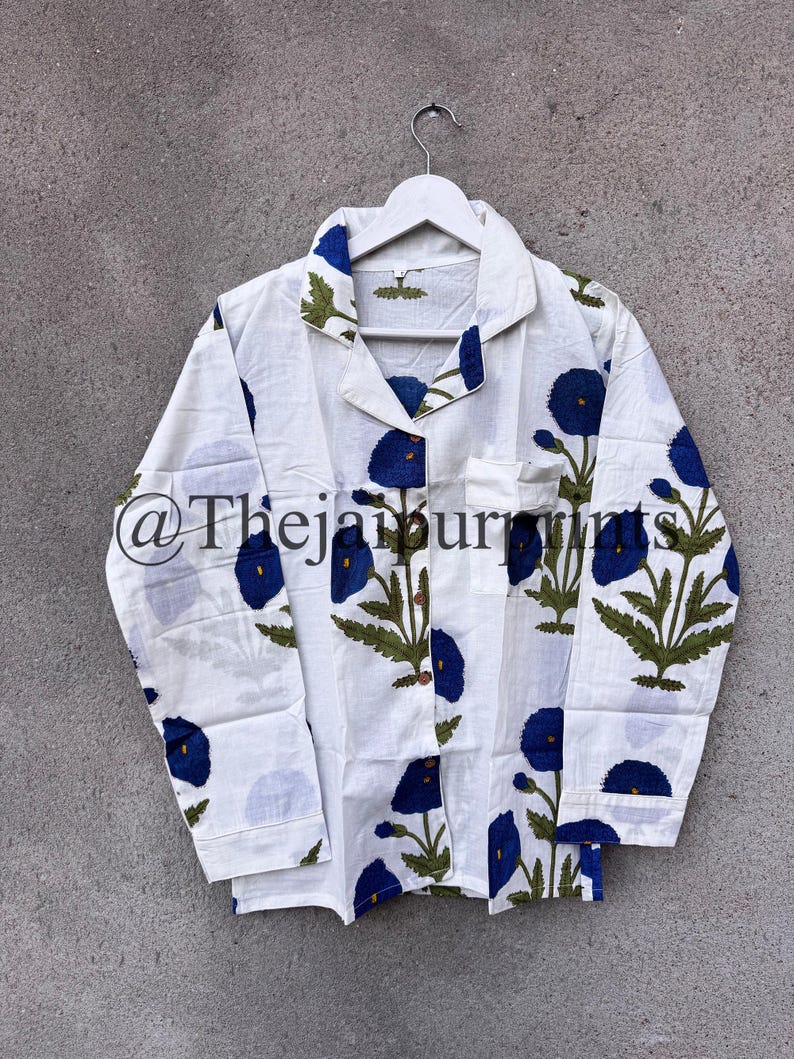 May include: White pajama top with a blue floral pattern. The long-sleeved shirt has a collared neckline and a pocket on the chest. The floral design features blue flowers and green leaves. The shirt is hanging on a white hanger.