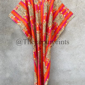 May include: A vibrant orange caftan with a tiger stripe pattern in black and tan, accented by pink stripes. The garment is hanging on a white hanger against a neutral background. The caftan features a V-neck and a drawstring.