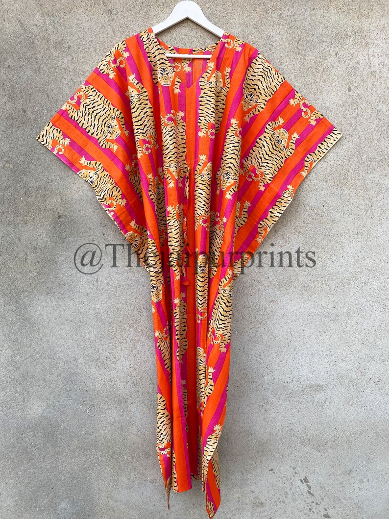 May include: A vibrant orange caftan with a tiger stripe pattern in black and tan, accented by pink stripes. The garment is hanging on a white hanger against a neutral background. The caftan features a V-neck and a drawstring.