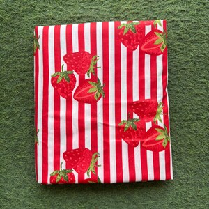 May include: Folded fabric with a red and white striped pattern. The fabric is decorated with red strawberries with green stems and leaves. The strawberries are whole and sliced, creating a playful design.