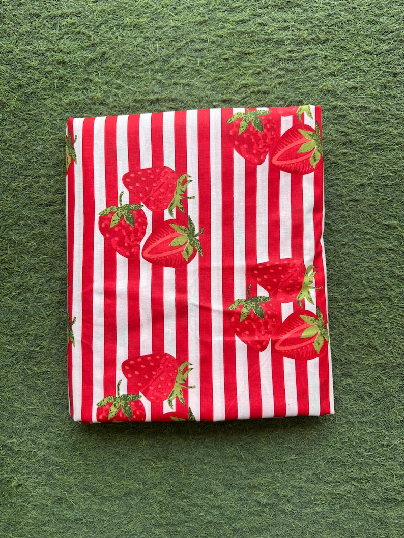 May include: Folded fabric with a red and white striped pattern. The fabric is decorated with red strawberries with green stems and leaves. The strawberries are whole and sliced, creating a playful design.