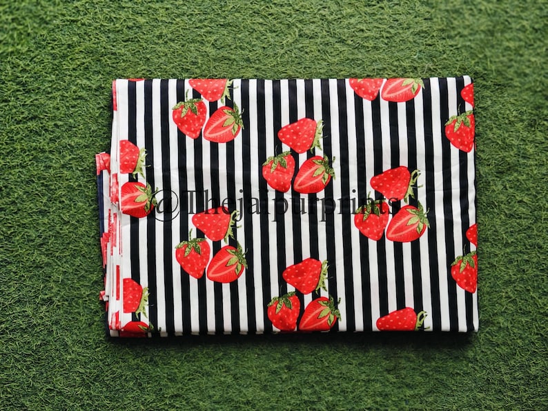 May include: Folded fabric with a black and white striped pattern. The fabric is decorated with red strawberries with green leaves. The text "@thejaprints" is visible on the fabric.