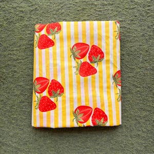 May include: Folded fabric with a yellow and white striped pattern, adorned with red strawberries. The strawberries have green stems and leaves. The fabric is likely cotton or a similar material, suitable for various sewing projects.