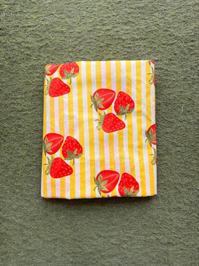 May include: Folded fabric with a yellow and white striped pattern, adorned with red strawberries. The strawberries have green stems and leaves. The fabric is likely cotton or a similar material, suitable for various sewing projects.