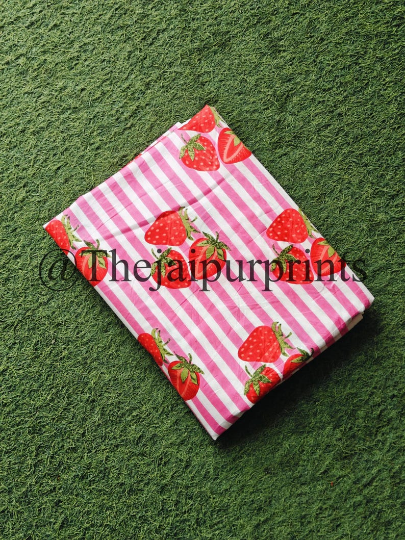 May include: A folded fabric with a pink and white striped pattern, adorned with illustrations of red strawberries. The fabric has the text "@thejaipurprints" printed on it.