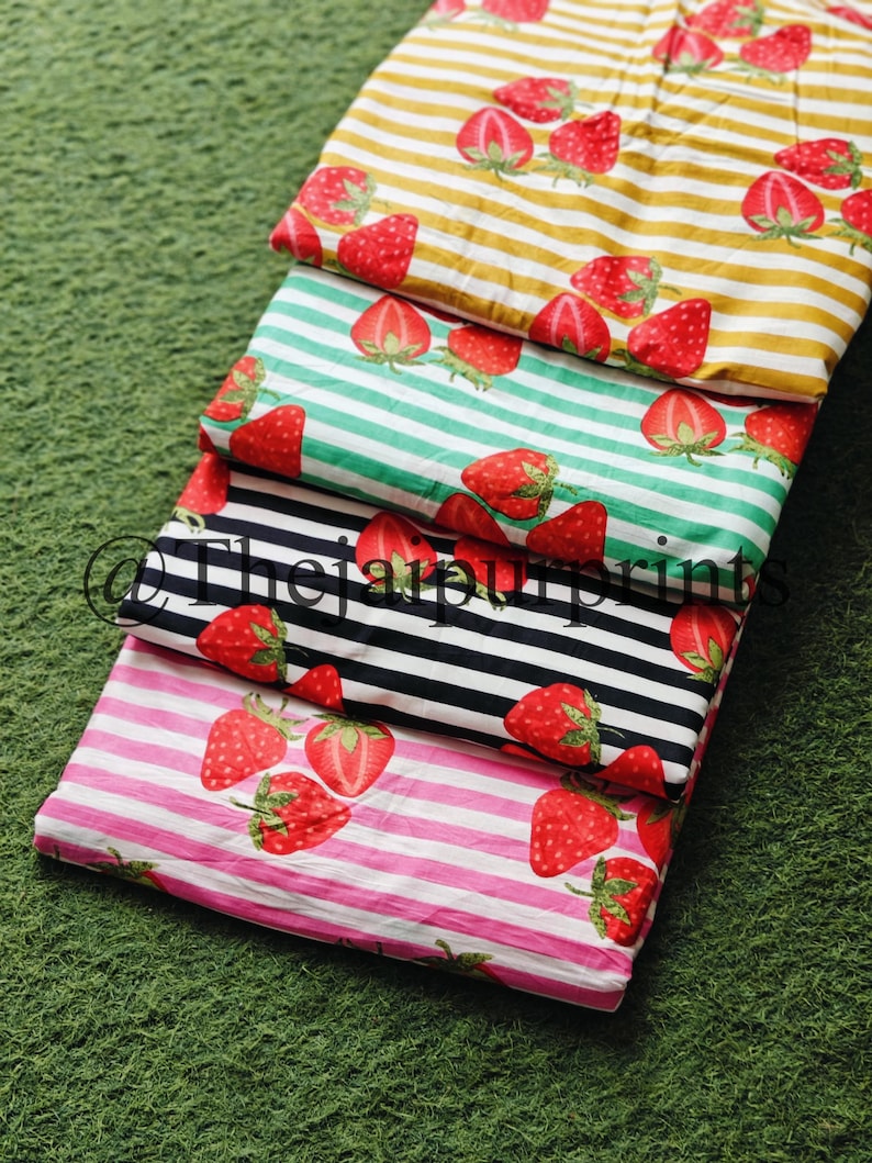 May include: Four fabric samples with a strawberry pattern. The fabrics feature red strawberries on striped backgrounds of yellow, green, black, and pink. The fabrics are folded and stacked on a green surface.