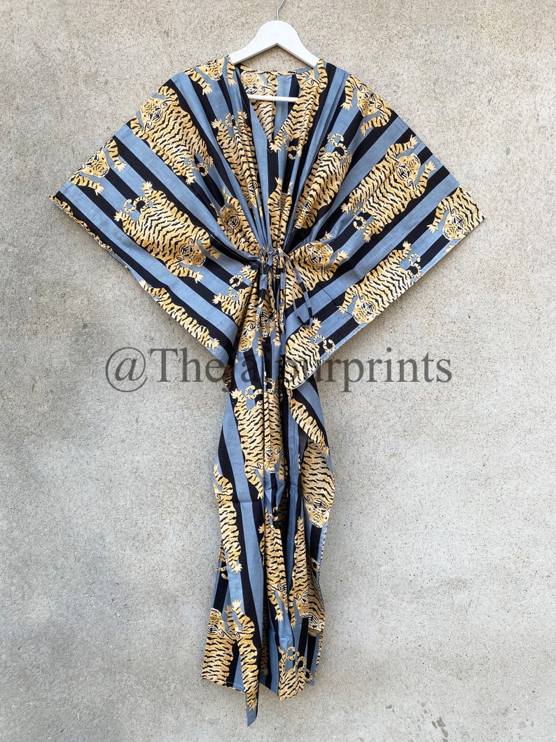 May include: A long, flowing kaftan dress with a V-neck, hanging on a white hanger. The dress features a repeating pattern of gold tigers on black and light blue striped fabric. The kaftan is tied at the waist with a matching fabric tie.