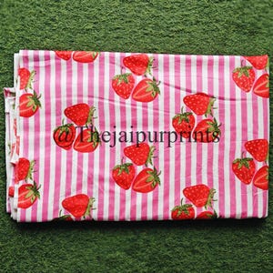 May include: A folded fabric with a repeating pattern of red strawberries and green leaves on a pink and white striped background. The text "@thejaipurprints" is visible on the fabric.