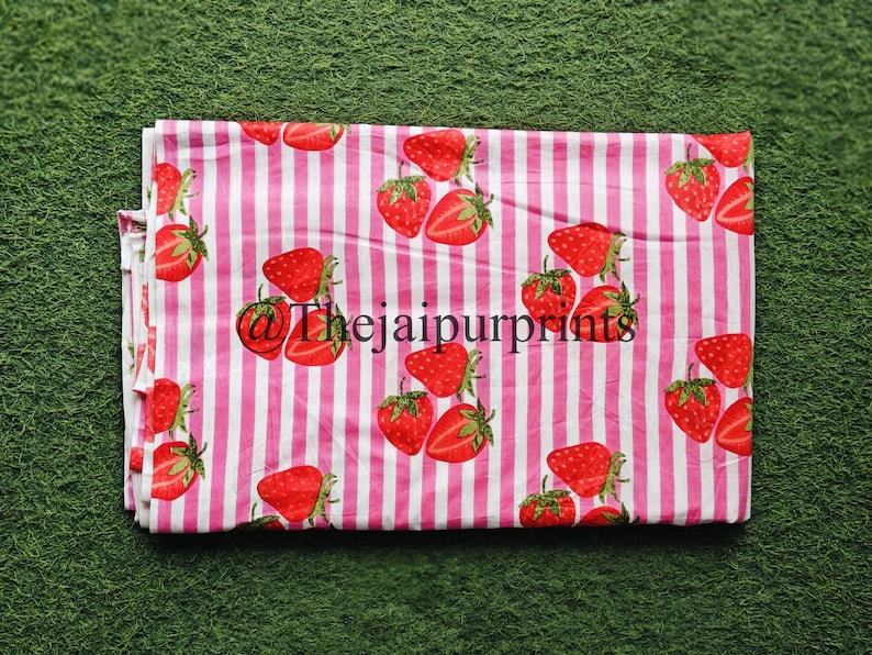 May include: A folded fabric with a repeating pattern of red strawberries and green leaves on a pink and white striped background. The text "@thejaipurprints" is visible on the fabric.