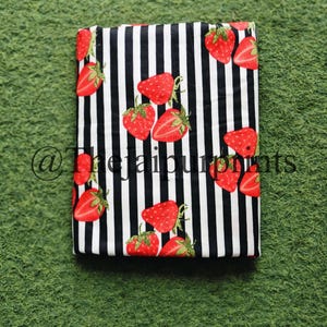 May include: Folded fabric with a black and white striped pattern, adorned with vibrant red strawberries. The strawberries have green stems and seeds. The fabric is rectangular and appears to be cotton or a similar material.