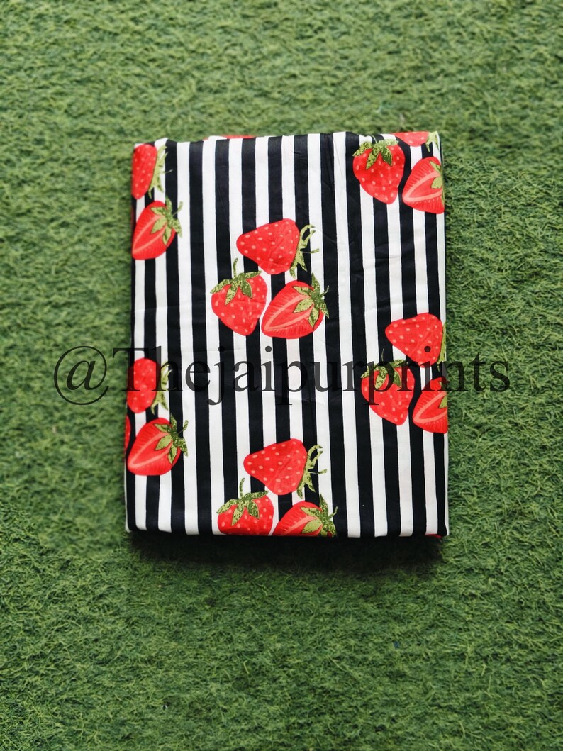 May include: Folded fabric with a black and white striped pattern, adorned with vibrant red strawberries. The strawberries have green stems and seeds. The fabric is rectangular and appears to be cotton or a similar material.