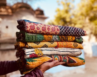 May include: A stack of colorful, patterned quilts held in a person's hands. The quilts feature various designs in red, blue, green, and yellow. The background is blurred, with a hint of a building and a blue sky.
