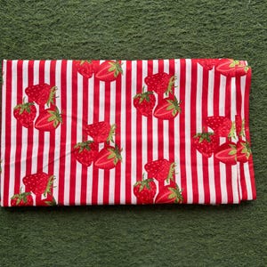 May include: Folded fabric with a red and white striped pattern. The fabric is decorated with illustrations of strawberries. The strawberries are red with green leaves. The fabric is folded and lies on a green surface.