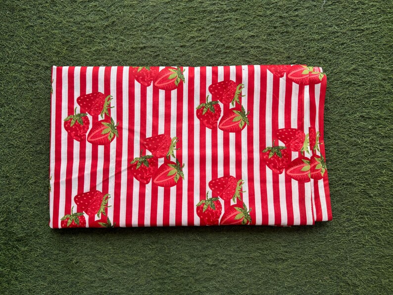 May include: Folded fabric with a red and white striped pattern. The fabric is decorated with illustrations of strawberries. The strawberries are red with green leaves. The fabric is folded and lies on a green surface.