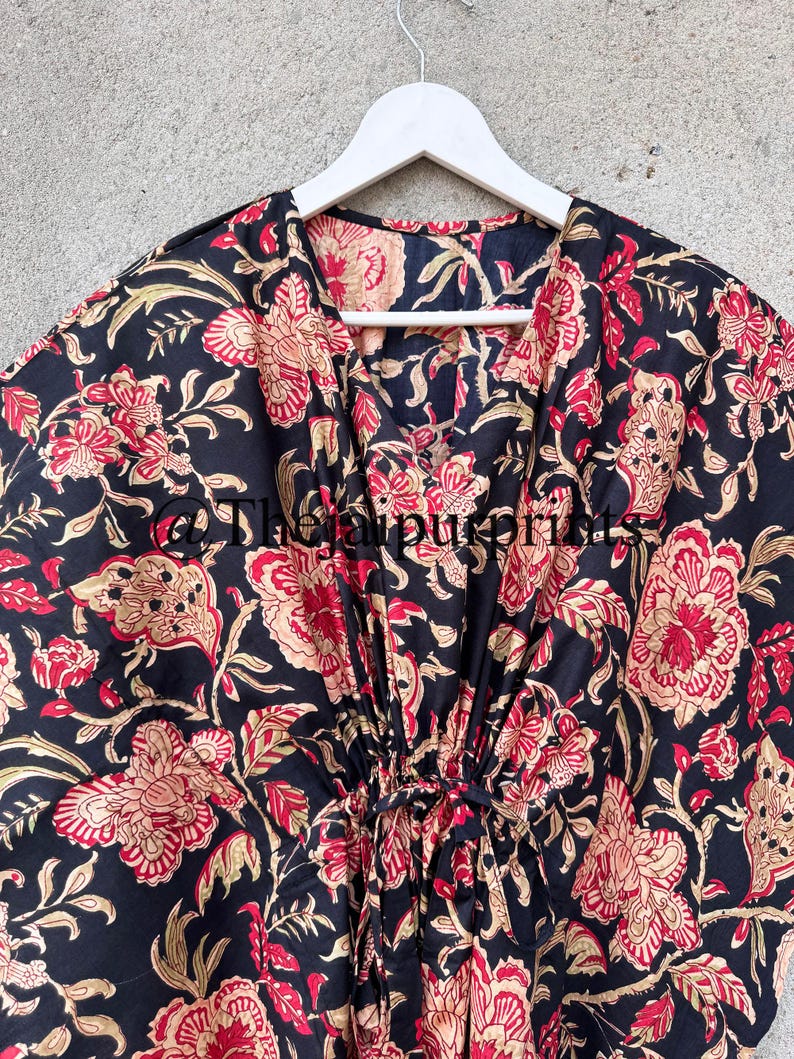 May include: Black cotton kaftan with a floral print in shades of red, gold, and cream. The kaftan has a V-neckline and a tie at the waist.