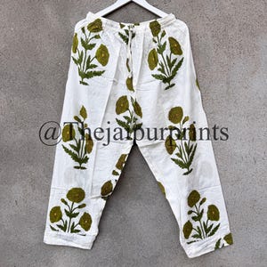 May include: White pants with a green floral pattern. The pants are hanging on a white hanger against a gray background. The text "@thejaipurprints" is visible on the pants.