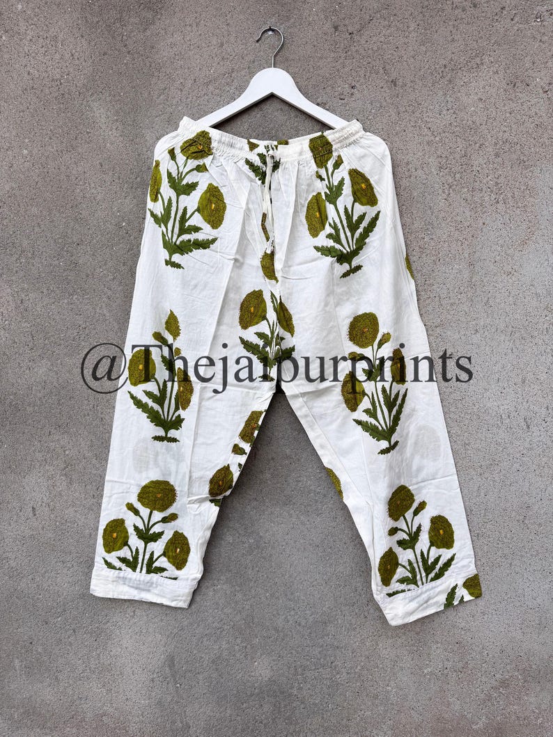 May include: White pants with a green floral pattern. The pants are hanging on a white hanger against a gray background. The text "@thejaipurprints" is visible on the pants.