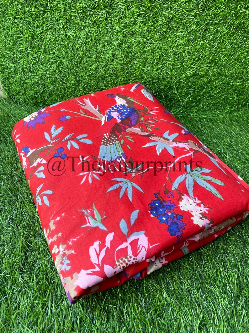 Op de afbeelding: Rode stof met een bloemen- en vogelprint. De stof heeft een herhalend patroon van roze bloemen, groene bladeren en blauwe vogels.