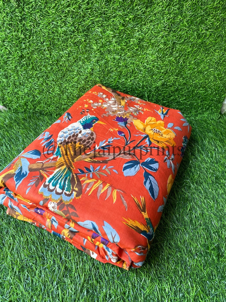 Op de afbeelding: Rode stof met een bloemen- en vogelprint. De stof heeft een grote vogel met groene, blauwe en bruine veren. De achtergrond is fel oranje met gele, witte en paarse bloemen.