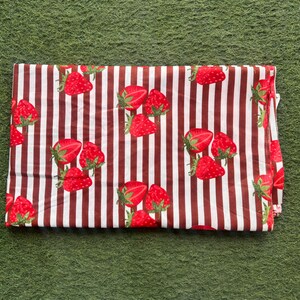 May include: Folded fabric with a repeating pattern of red strawberries and brown vertical stripes on a white background. The strawberries have green stems and leaves. The fabric is folded and appears to be made of cotton or a similar material.