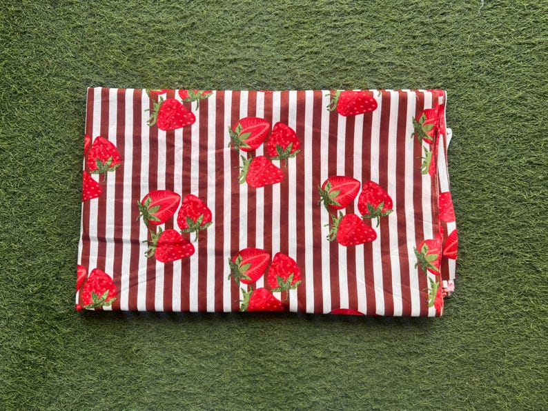 May include: Folded fabric with a repeating pattern of red strawberries and brown vertical stripes on a white background. The strawberries have green stems and leaves. The fabric is folded and appears to be made of cotton or a similar material.
