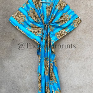 May include: A blue and gold tiger print kaftan dress. The dress features a striped pattern with tiger illustrations. The kaftan has a drawstring waist and is hanging on a white hanger against a neutral background.
