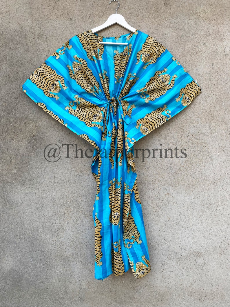May include: A blue and gold tiger print kaftan dress. The dress features a striped pattern with tiger illustrations. The kaftan has a drawstring waist and is hanging on a white hanger against a neutral background.