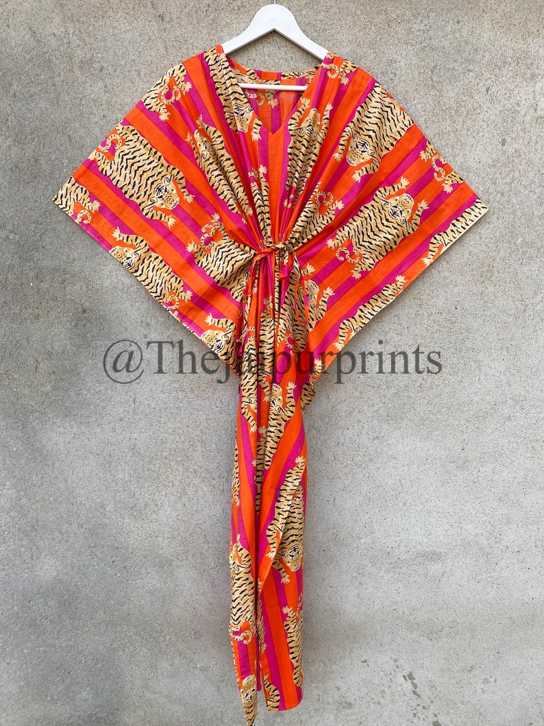 May include: An orange and pink patterned kaftan dress with a tiger print design. The dress has a V-neck and a tie at the waist. The fabric is lightweight and appears to be cotton. The Jaipur Prints is printed on the bottom left.