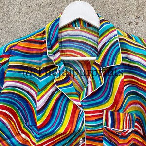 May include: A colorful striped shirt with a white collar and a pocket on the left side. The shirt has a wavy pattern and is made of a soft, silky fabric. The shirt is hanging on a white hanger.
