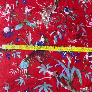 Op de afbeelding: Rode katoenen stof met een bloemen- en vogelprint. De stof wordt gemeten met een gele meetlint.