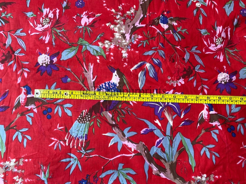 Op de afbeelding: Rode katoenen stof met een bloemen- en vogelprint. De stof wordt gemeten met een gele meetlint.