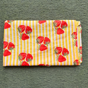 May include: Folded fabric featuring a repeating pattern of red strawberries with green stems on a yellow and white striped background. The fabric is folded neatly, showcasing the design.